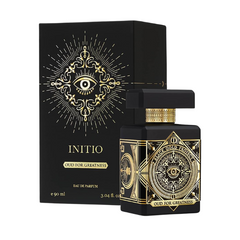 Product photograph of Initio Oud for Greatness Eau de Parfum on a white background showcasing a black bottle with gold geometric designs and a central all-seeing eye, next to its matching box featuring sunburst rays and Black Gold Project branding