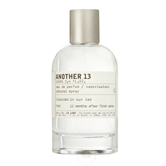 Product photo of Le Labo Another 13 Eau de Parfum, a minimalist 100ml clear glass bottle with a brushed silver cap and white label detailing its New York and Paris origins, with text 'Compounded in our lab' and '12 months after first spray'