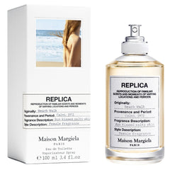 Maison Margiela Replica Beach Walk Eau de Toilette clear glass bottle and white packaging box, featuring a beach scene with a woman on a sandy shore by the blue ocean, detailed with labels citing Calvi 1972 and a sun-kissed salty skin fragrance