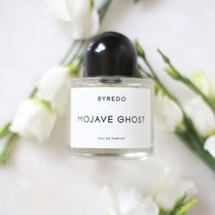 Product photo of a Byredo Mojave Ghost Eau de Parfum bottle with a sleek black cap and white label, elegantly displayed on a clean white surface adorned with delicate white flowers and green foliage
