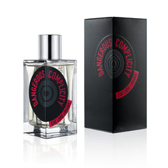 Product photography by Etat Libre d'Orange featuring the Dangerous Complicity fragrance in a clear glass bottle with a silver cap and a black circular label with red text, alongside its matching black embossed box, all on a white reflective surface