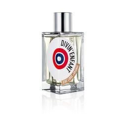 Photography by Etat Libre d'Orange depicting the DIVIN'ENFANT perfume bottle on a white background, featuring a clear glass body, shiny silver cap, pale yellow liquid, and a white label with prominent black text, a striking red-white-blue target emblem, and a brown Etat Libre d'Orange banner