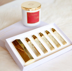 Photo of a Maison Francis Kurkdjian fragrance set including a gold geometric bottle and five Baccarat Rouge 540 travel sprays in a white box, alongside a white candle with a red label and gold lid, all on a soft white surface