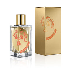 Product photo of a La Fin Du Monde perfume bottle by Etat Libre d'Orange featuring a golden cap and a golden circular label with orange text and a mushroom cloud graphic, displayed next to its matching matte gold embossed box on a reflective white surface