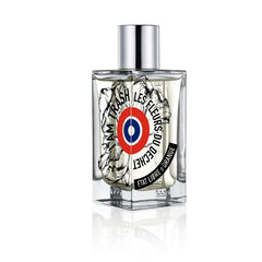Etat Libre d'Orange perfume bottle, a clear square glass bottle with a silver cap, features a unique label displaying I Am Trash Les Fleurs Du Déchet text, a red white and blue target symbol, and intricate black and white floral illustrations, reflected on a white background