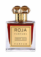 Product photograph of a Roja Parfums Amber Aoud perfume bottle featuring a clear glass bottle with rich amber liquid, a gold label, and a distinctive gold cap topped with two faceted amber-brown stones