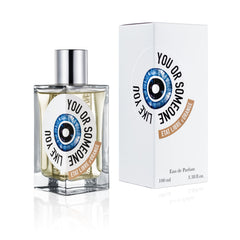 Product photography of the Etat Libre d'Orange You Or Someone Like You Eau de Parfum, showing a clear glass bottle with a silver cap and light amber liquid, featuring a distinctive circular blue eye label with black text and an orange-brown brand banner, displayed next to its matching white box