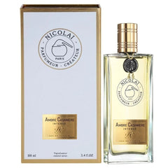 Photography of Nicolai Parfumeur Créateur's Ambre Cashmere Intense Eau de Parfum, showing a clear rectangular glass bottle with a ribbed gold cap and black 'N' wax seal next to its white and gold box featuring the brand's Paris flask logo