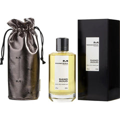 Product photography by Mancera Paris of the Sand Aoud Eau de Parfum set, featuring a clear bottle with pale yellow liquid, a metallic gunmetal drawstring pouch, and a matte black box displaying the brand's logo